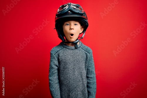 Young little caucasian kid wearing vintage biker motorcycle helmet and googles over red background afraid and shocked with surprise expression, fear and excited face.