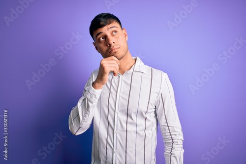 Young handsome hispanic man wearing elegant business shirt standing over purple background with hand on chin thinking about question, pensive expression. Smiling with thoughtful face. Doubt concept.