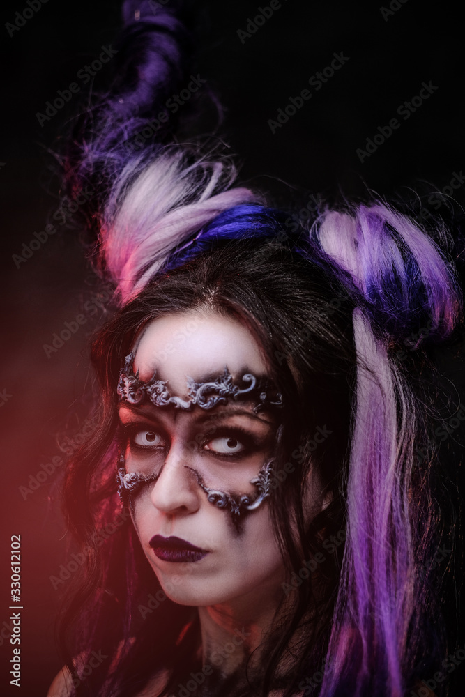 Fototapeta premium Portrait of a demonic girl wearing scary fantasy make up and violet horns posing in a dark studio with red light on a grey background and looking spooky. Close up view.