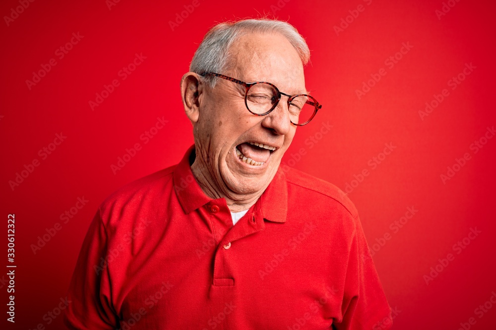 Grey haired senior man wearing glasses and casual t-shirt over red background winking looking at the camera with sexy expression, cheerful and happy face. Grey haired senior man wearing glasses and casual t-shirt over red background winking looking at the camera with sexy expression, cheerful and happy face.