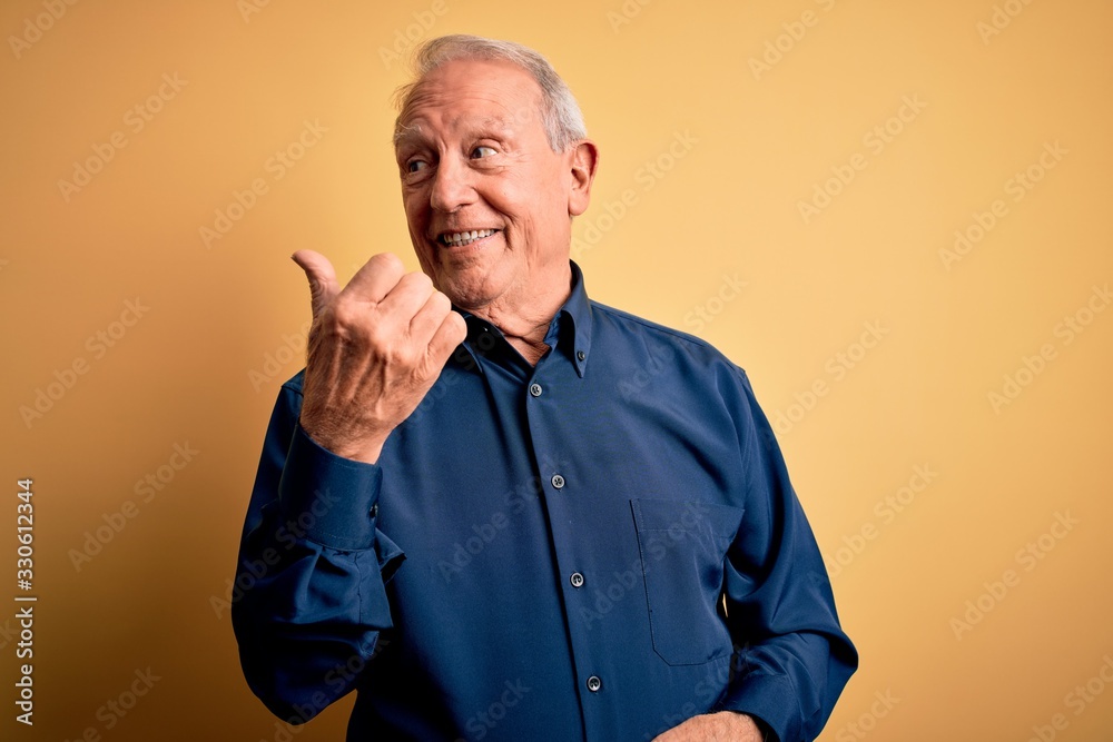 Grey haired senior man wearing casual blue shirt standing over yellow background smiling with happy face looking and pointing to the side with thumb up. Grey haired senior man wearing casual blue shirt standing over yellow background smiling with happy face looking and pointing to the side with thumb up.
