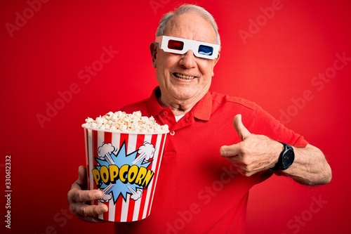 Grey haired senior man wearing 3d movie glasses and eating popcorn over red background happy with big smile doing ok sign, thumb up with fingers, excellent sign