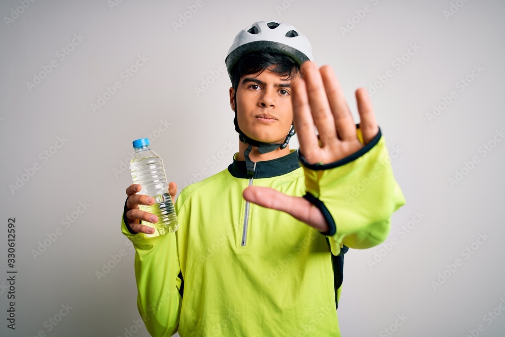 Young handsome cyclist man wearing security bike helmet drinking bottle of water with open hand doing stop sign with serious and confident expression, defense gesture