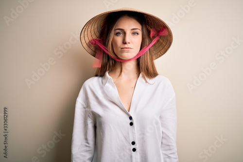Young beautiful redhead woman wearing asian traditional conical hat over white background with serious expression on face. Simple and natural looking at the camera.