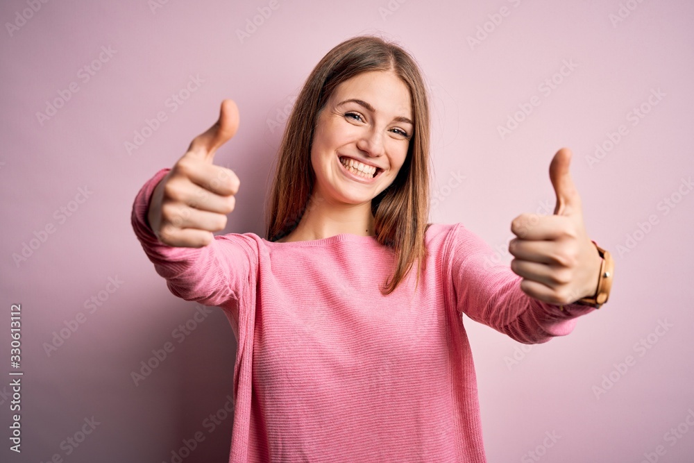 Young beautiful redhead woman wearing casual sweater over isolated blue background approving doing positive gesture with hand, thumbs up smiling and happy for success. Winner gesture.