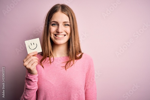 Young beautiful redhead woman holding reminder paper with smile emoji message with a happy face standing and smiling with a confident smile showing teeth