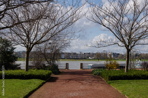 Diminishing perspective view of red clay path between Rheingärtchen garden with background of Rhine river and overcast sky.