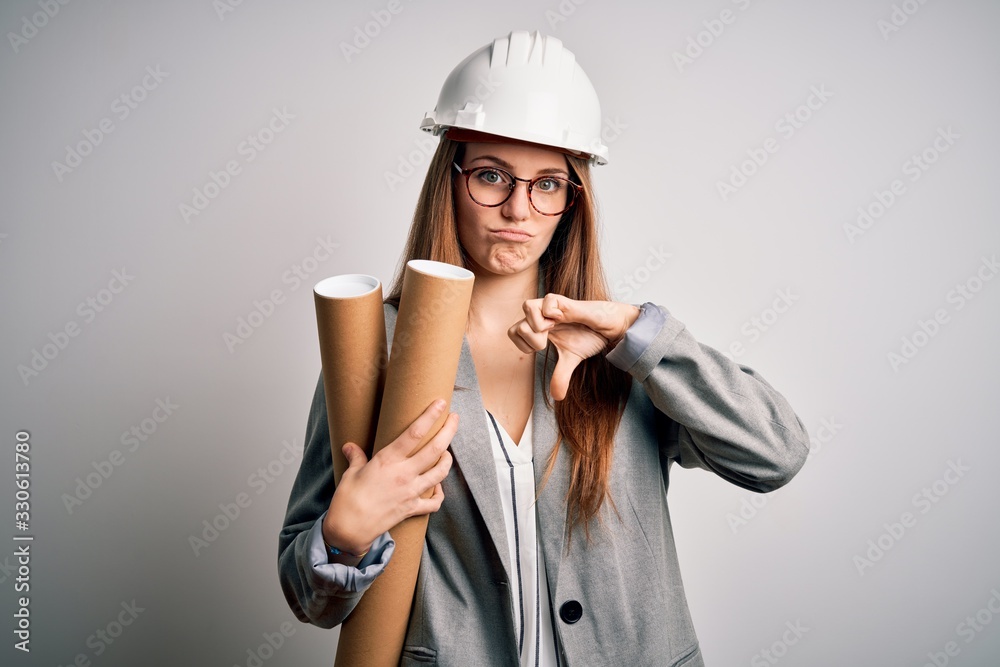 Young beautiful redhead architect woman wearing glasses and safety helmet holding blueprints with angry face, negative sign showing dislike with thumbs down, rejection concept