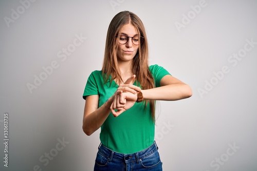 Young beautiful redhead woman wearing casual green t-shirt and glasses over white background Checking the time on wrist watch  relaxed and confident