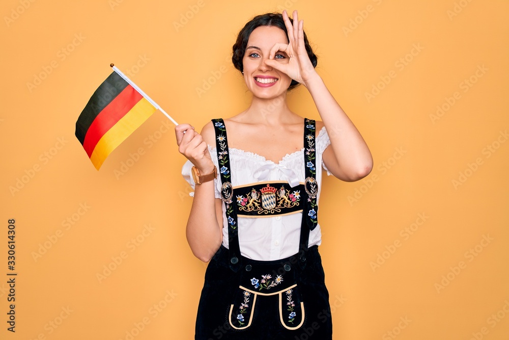 Young patriotic german woman with blue eyes wearing octoberfest dress holding germany flag with happy face smiling doing ok sign with hand on eye looking through fingers Young patriotic german woman with blue eyes wearing octoberfest dress holding germany flag with happy face smiling doing ok sign with hand on eye looking through fingers