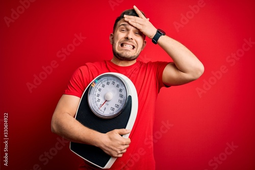Young fitness man with blue eyes holding scale dieting for healthy weight over red background stressed with hand on head, shocked with shame and surprise face, angry and frustrated. Fear and upset