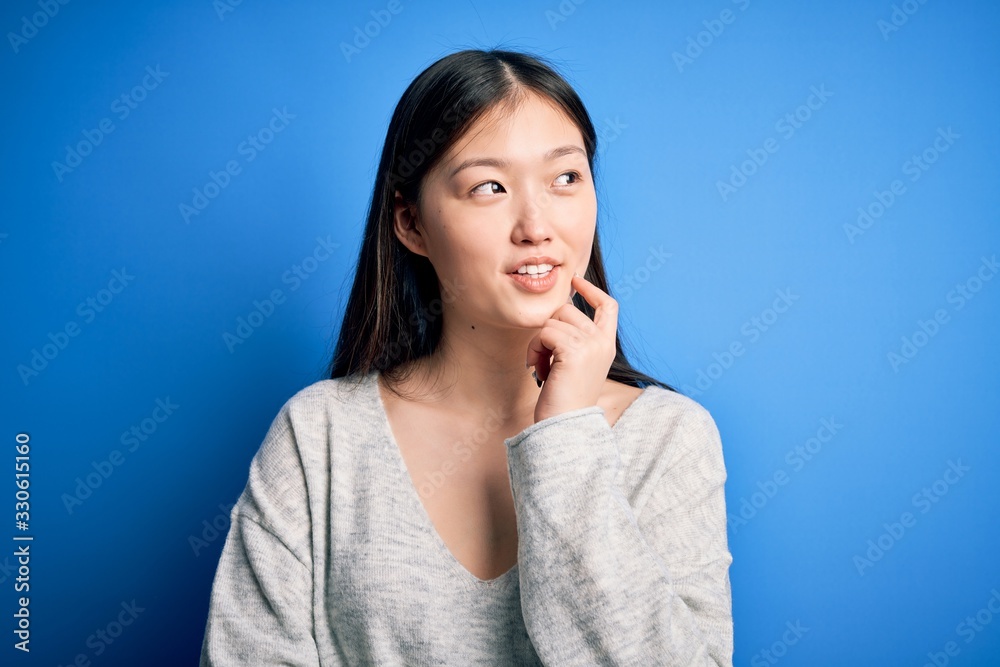 Young beautiful asian woman wearing casual sweater standing over blue isolated background with hand on chin thinking about question, pensive expression. Smiling and thoughtful face. Doubt concept. Young beautiful asian woman wearing casual sweater standing over blue isolated background with hand on chin thinking about question, pensive expression. Smiling and thoughtful face. Doubt concept.