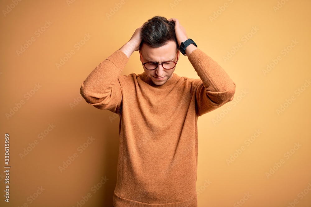 Young handsome caucasian man wearing glasses and casual winter sweater over yellow background suffering from headache desperate and stressed because pain and migraine. Hands on head. Young handsome caucasian man wearing glasses and casual winter sweater over yellow background suffering from headache desperate and stressed because pain and migraine. Hands on head.
