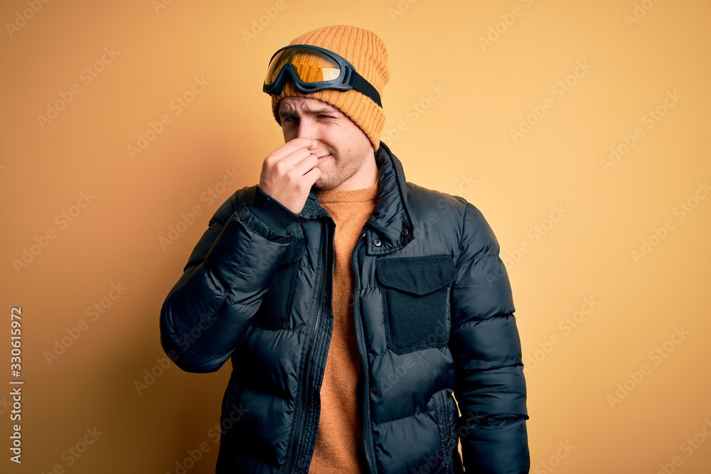 Young handsome caucasian man wearing hat, coat and ski glasses for winter and snow weather smelling something stinky and disgusting, intolerable smell, holding breath with fingers on nose. Bad smell Young handsome caucasian man wearing hat, coat and ski glasses for winter and snow weather smelling something stinky and disgusting, intolerable smell, holding breath with fingers on nose. Bad smell