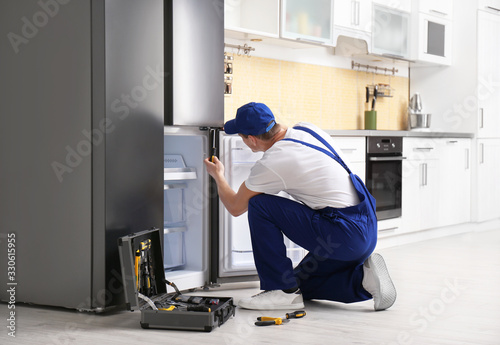 Male technician with screwdriver repairing refrigerator in kitchen photo
