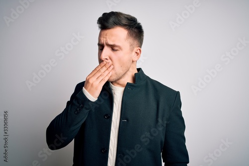 Young handsome business mas wearing elegant winter coat standing over isolated background bored yawning tired covering mouth with hand. Restless and sleepiness.