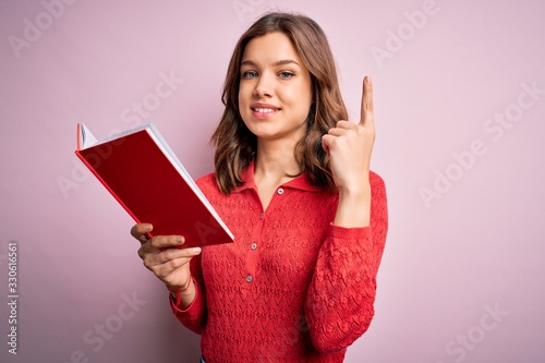 Young blonde student girl reading a book over pink  isolated background surprised with an idea or question pointing finger with happy face  number one