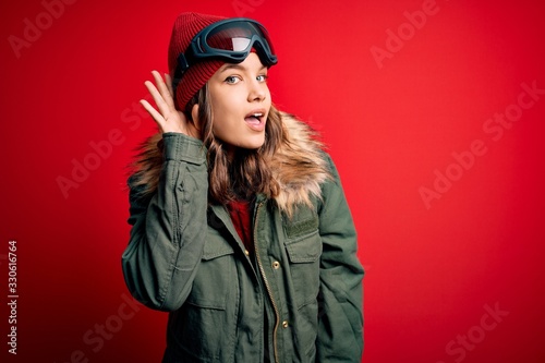Young blonde girl wearing ski glasses and winter coat for ski weather over red background smiling with hand over ear listening an hearing to rumor or gossip. Deafness concept.