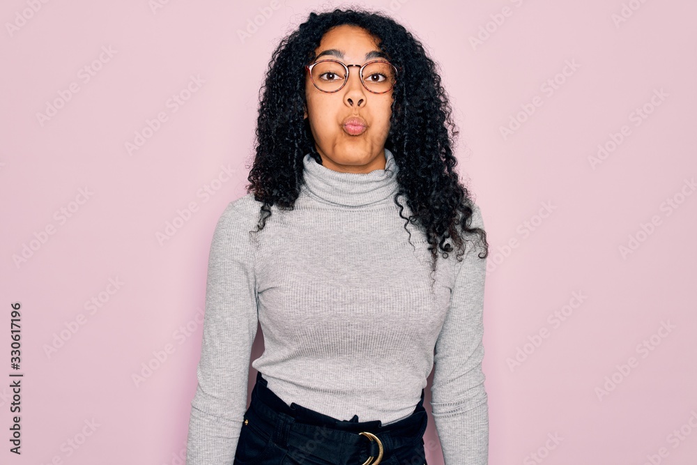 Young african american woman wearing turtleneck sweater and glasses over pink background puffing cheeks with funny face. Mouth inflated with air, crazy expression. Young african american woman wearing turtleneck sweater and glasses over pink background puffing cheeks with funny face. Mouth inflated with air, crazy expression.