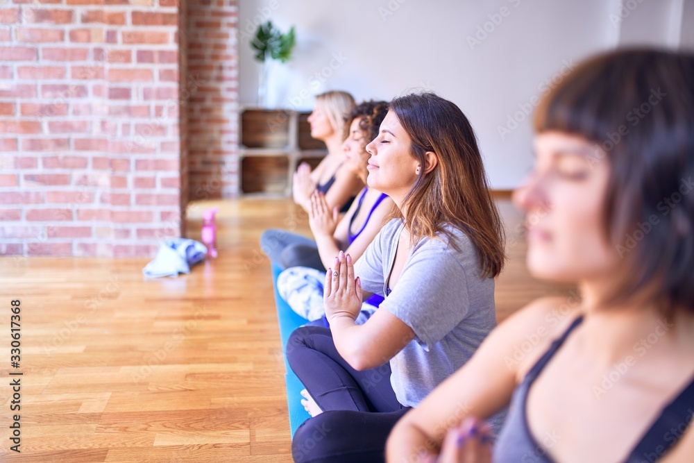 Young beautiful group of sportswomen smiling happy practicing yoga. Sitting doing prayer pose at gym Young beautiful group of sportswomen smiling happy practicing yoga. Sitting doing prayer pose at gym