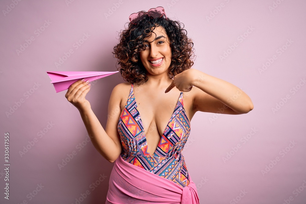 Young beautiful curly arab woman on vacation wearing swimsuit holding pink paper airplane with surprise face pointing finger to himself Young beautiful curly arab woman on vacation wearing swimsuit holding pink paper airplane with surprise face pointing finger to himself