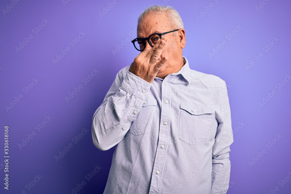 Middle age handsome hoary man wearing striped shirt and glasses over purple background smelling something stinky and disgusting, intolerable smell, holding breath with fingers on nose. Bad smell Middle age handsome hoary man wearing striped shirt and glasses over purple background smelling something stinky and disgusting, intolerable smell, holding breath with fingers on nose. Bad smell