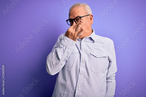 Middle age handsome hoary man wearing striped shirt and glasses over purple background smelling something stinky and disgusting, intolerable smell, holding breath with fingers on nose. Bad smell