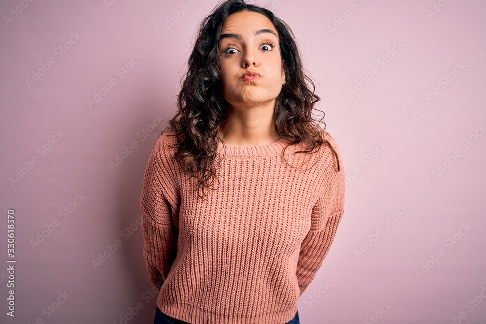 Obraz premium Young beautiful woman with curly hair wearing casual sweater over isolated pink background puffing cheeks with funny face. Mouth inflated with air, crazy expression.