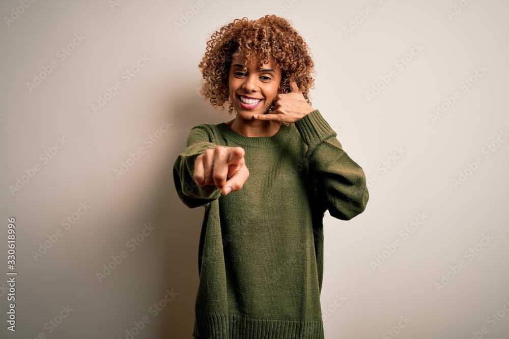 Beautiful african american woman with curly hair wearing casual sweater over white background smiling doing talking on the telephone gesture and pointing to you. Call me. Beautiful african american woman with curly hair wearing casual sweater over white background smiling doing talking on the telephone gesture and pointing to you. Call me.