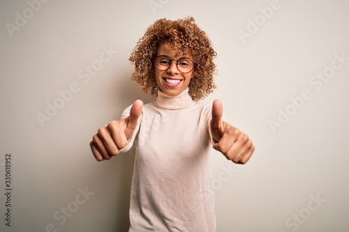 Young african american woman wearing turtleneck sweater and glasses over white background approving doing positive gesture with hand, thumbs up smiling and happy for success. Winner gesture.