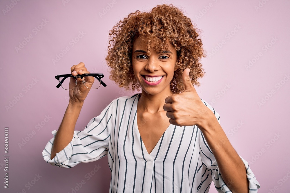Obraz premium African american optical woman with curly hair holding glasses over isolated pink background happy with big smile doing ok sign, thumb up with fingers, excellent sign