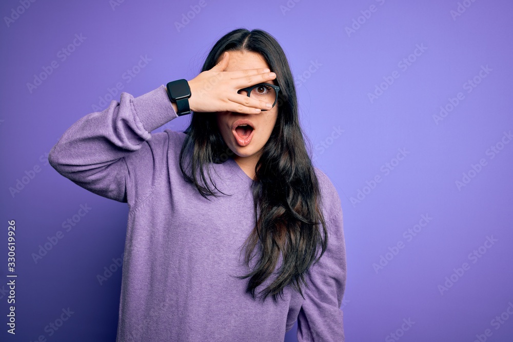 Young brunette woman wearing glasses over purple isolated background peeking in shock covering face and eyes with hand, looking through fingers with embarrassed expression. Young brunette woman wearing glasses over purple isolated background peeking in shock covering face and eyes with hand, looking through fingers with embarrassed expression.