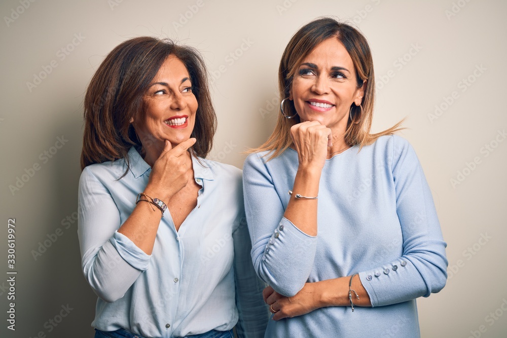 Middle age beautiful couple of sisters standing over isolated white background with hand on chin thinking about question, pensive expression. Smiling with thoughtful face. Doubt concept.