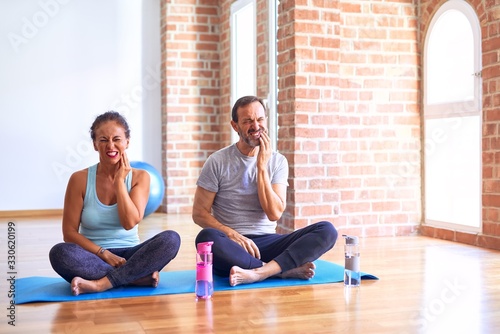 Middle age sporty couple sitting on mat doing stretching yoga exercise at gym touching mouth with hand with painful expression because of toothache or dental illness on teeth. Dentist concept.