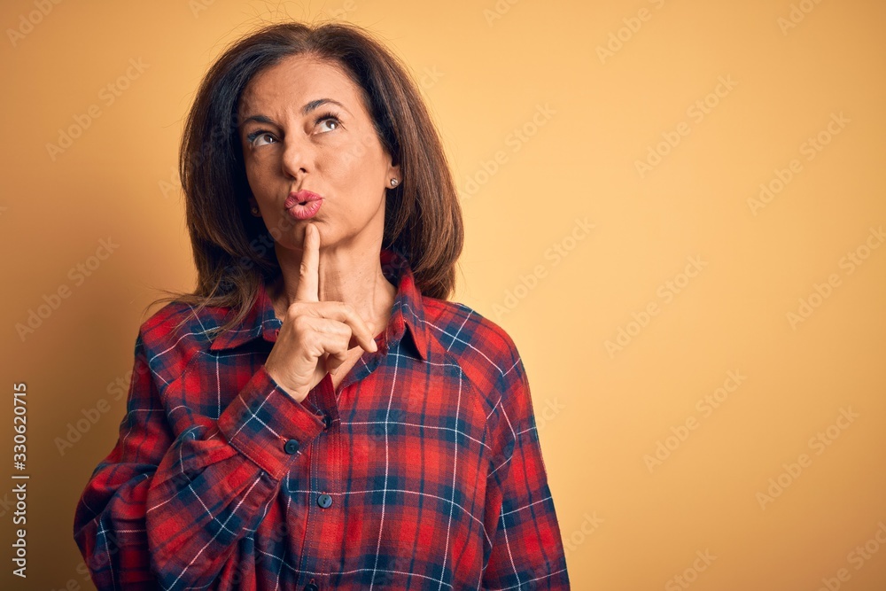 Middle age beautiful woman wearing casual shirt standing over isolated yellow background Thinking concentrated about doubt with finger on chin and looking up wondering Middle age beautiful woman wearing casual shirt standing over isolated yellow background Thinking concentrated about doubt with finger on chin and looking up wondering