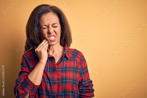 Middle age beautiful woman wearing casual shirt standing over isolated yellow background touching mouth with hand with painful expression because of toothache or dental illness on teeth. Dentist