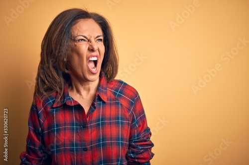 Middle age beautiful woman wearing casual shirt standing over isolated yellow background angry and mad screaming frustrated and furious, shouting with anger. Rage and aggressive concept.