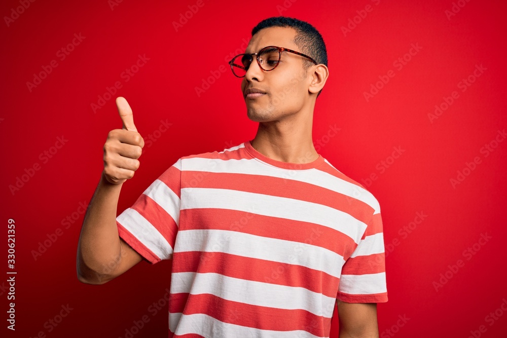 Naklejka premium Young handsome african american man wearing casual striped t-shirt and glasses Looking proud, smiling doing thumbs up gesture to the side