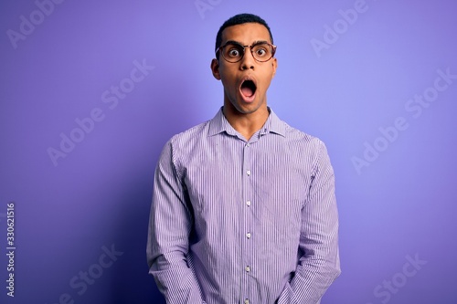 Handsome african american man wearing striped shirt and glasses over purple background afraid and shocked with surprise and amazed expression, fear and excited face.