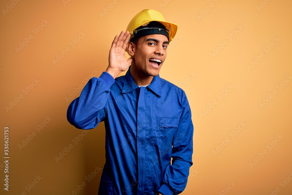 Young handsome african american worker man wearing blue uniform and security helmet smiling with hand over ear listening an hearing to rumor or gossip. Deafness concept.