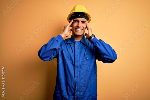 Young handsome african american worker man wearing blue uniform and security helmet covering ears with fingers with annoyed expression for the noise of loud music. Deaf concept.