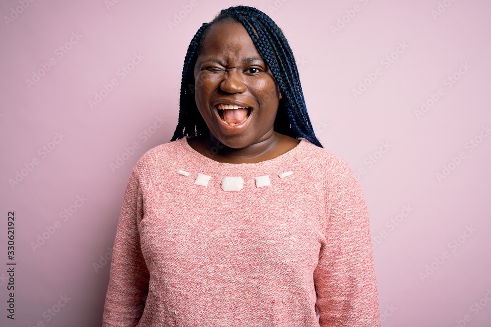African american plus size woman with braids wearing casual sweater over pink background winking looking at the camera with sexy expression, cheerful and happy face. African american plus size woman with braids wearing casual sweater over pink background winking looking at the camera with sexy expression, cheerful and happy face.