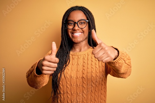 Young african american smart woman wearing glasses and casual sweater over yellow background approving doing positive gesture with hand, thumbs up smiling and happy for success. Winner gesture.