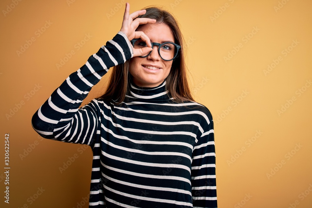 Beautiful woman with blue eyes wearing striped sweater and glasses over yellow background doing ok gesture with hand smiling, eye looking through fingers with happy face. Beautiful woman with blue eyes wearing striped sweater and glasses over yellow background doing ok gesture with hand smiling, eye looking through fingers with happy face.
