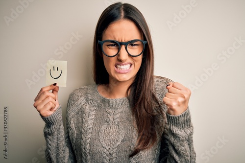 Fototapeta Naklejka Na Ścianę i Meble -  Young beautiful brunette woman wearing glasses holding paper with smile emoji annoyed and frustrated shouting with anger, crazy and yelling with raised hand, anger concept
