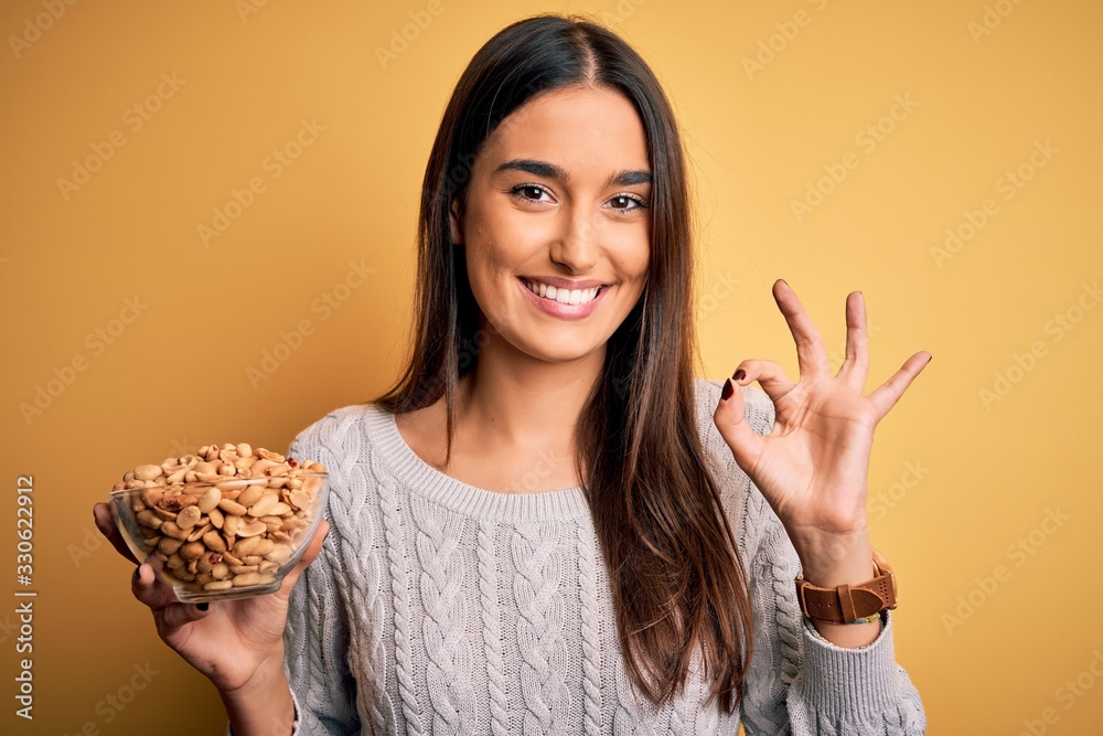 Young beautiful brunette woman holding bowl with peanuts over isolated yellow background doing ok sign with fingers, excellent symbol Young beautiful brunette woman holding bowl with peanuts over isolated yellow background doing ok sign with fingers, excellent symbol