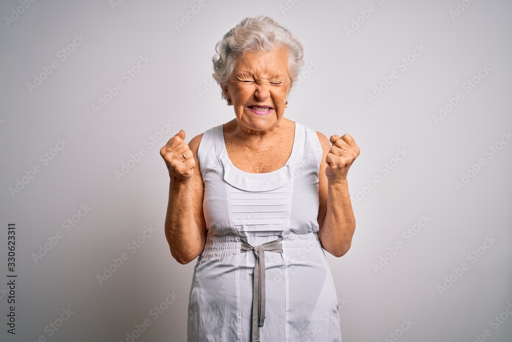 Senior beautiful grey-haired woman wearing casual summer dress over white background very happy and excited doing winner gesture with arms raised, smiling and screaming for success. Celebration Senior beautiful grey-haired woman wearing casual summer dress over white background very happy and excited doing winner gesture with arms raised, smiling and screaming for success. Celebration