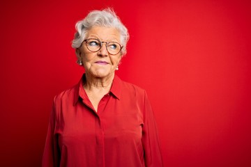 Senior beautiful grey-haired woman wearing casual shirt and glasses over red background smiling looking to the side and staring away thinking.