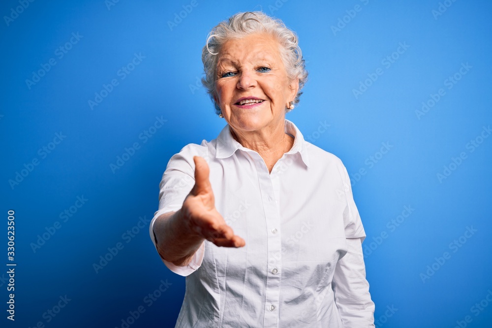 Senior beautiful woman wearing elegant shirt standing over isolated blue background smiling friendly offering handshake as greeting and welcoming. Successful business. Senior beautiful woman wearing elegant shirt standing over isolated blue background smiling friendly offering handshake as greeting and welcoming. Successful business.