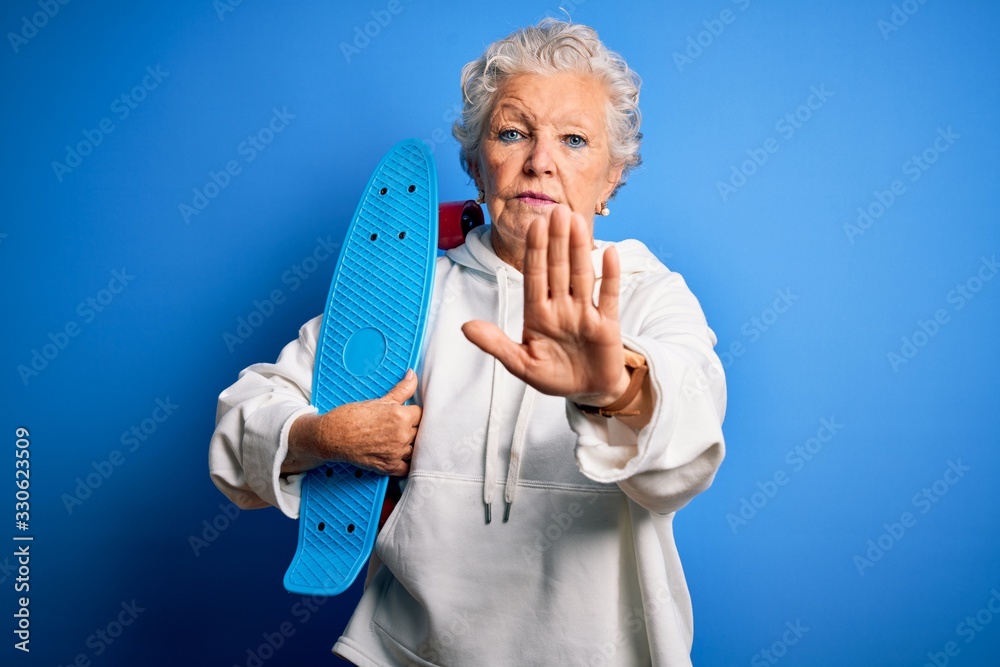Senior beautiful sporty woman holding skate standing over isolated blue background with open hand doing stop sign with serious and confident expression, defense gesture Senior beautiful sporty woman holding skate standing over isolated blue background with open hand doing stop sign with serious and confident expression, defense gesture